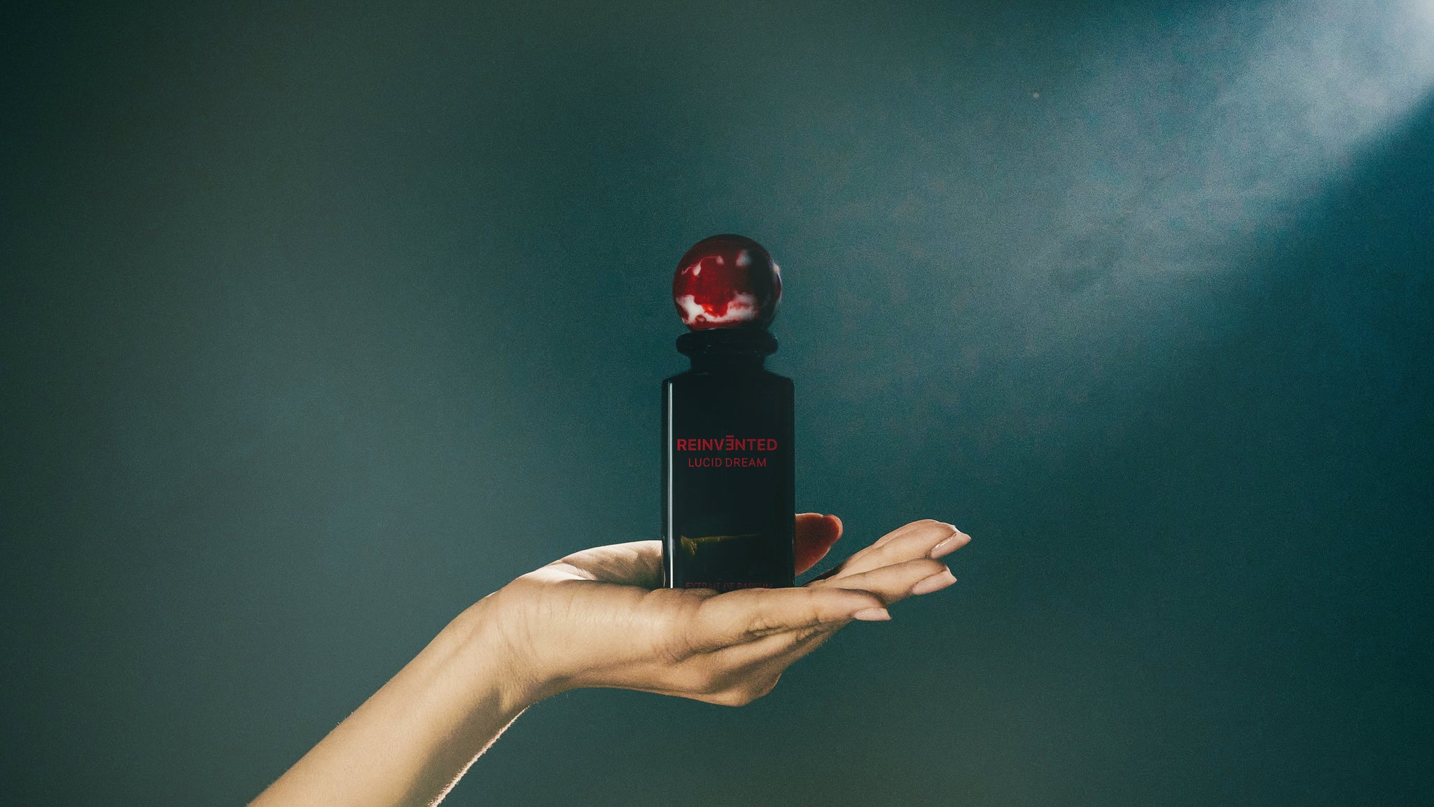 A hand with manicured nails holds a dark perfume bottle labeled REINVENTED LUCIDDREAM with a red, marbled spherical cap against a moody, softly lit background.
