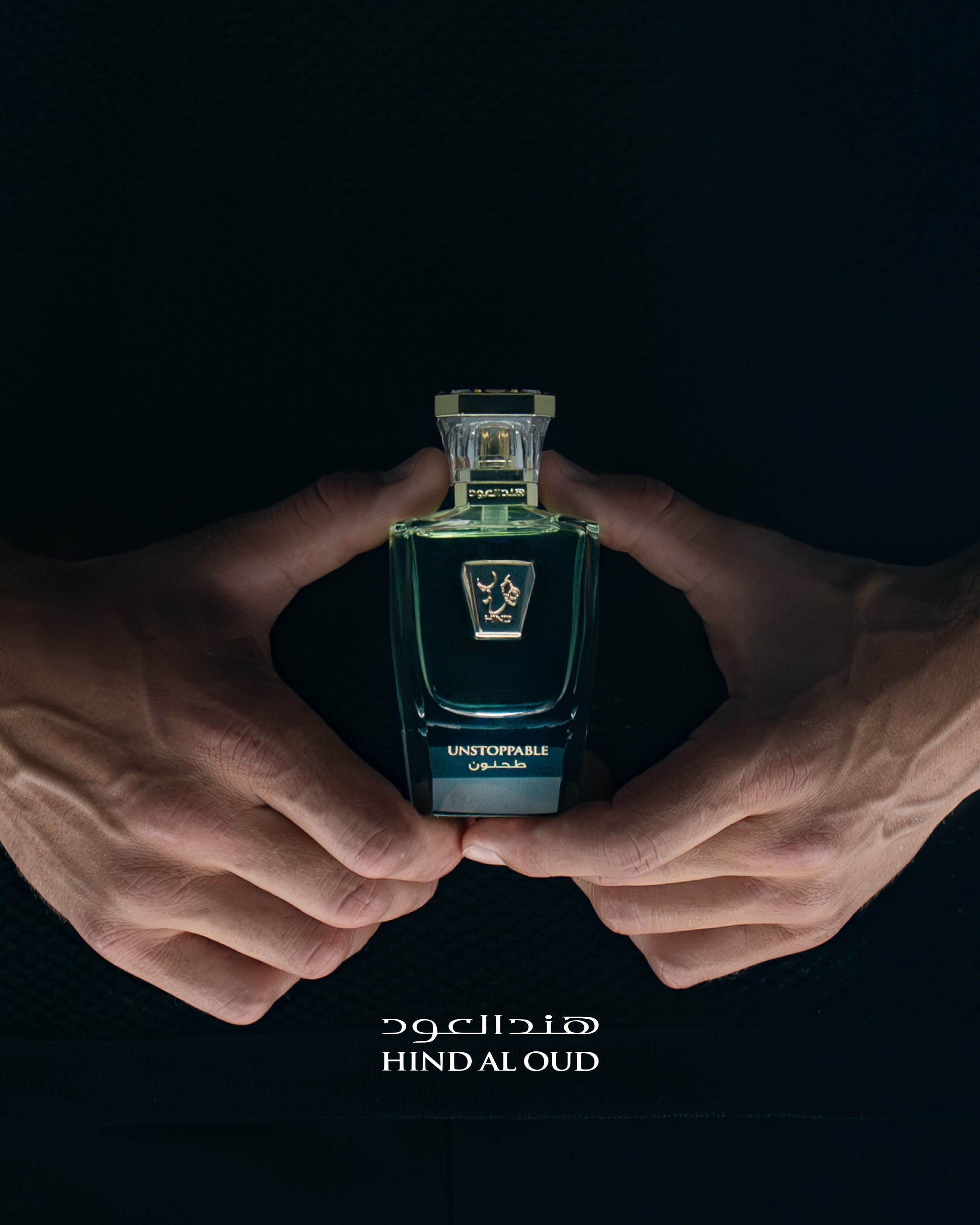 A persons hands holding a green bottle of perfume labeled UNSTOPPABLE by Hind Al Oud against a black background, with the Hind Al Oud logo and name in Arabic and English below.