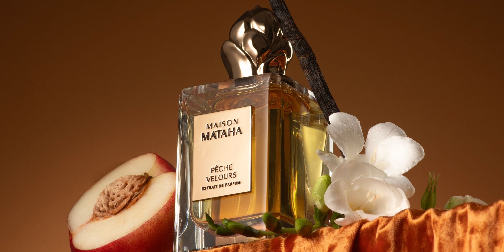 A bottle of Maison Mataha Pêche Velours perfume sits on orange velvet, surrounded by a peach half, a vanilla bean, and white flowers, set against a warm brown background.
