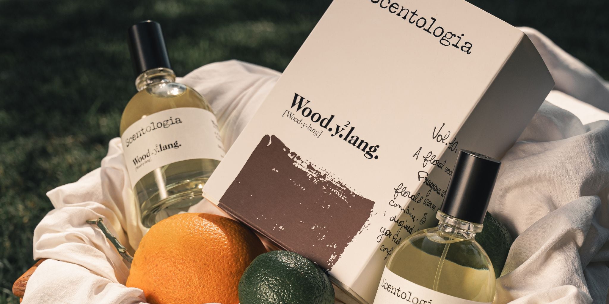 Two glass bottles and a box of Scentologia’s Wood Ylang perfume rest in a basket with an orange and two avocados on a white cloth, set outdoors on green grass.